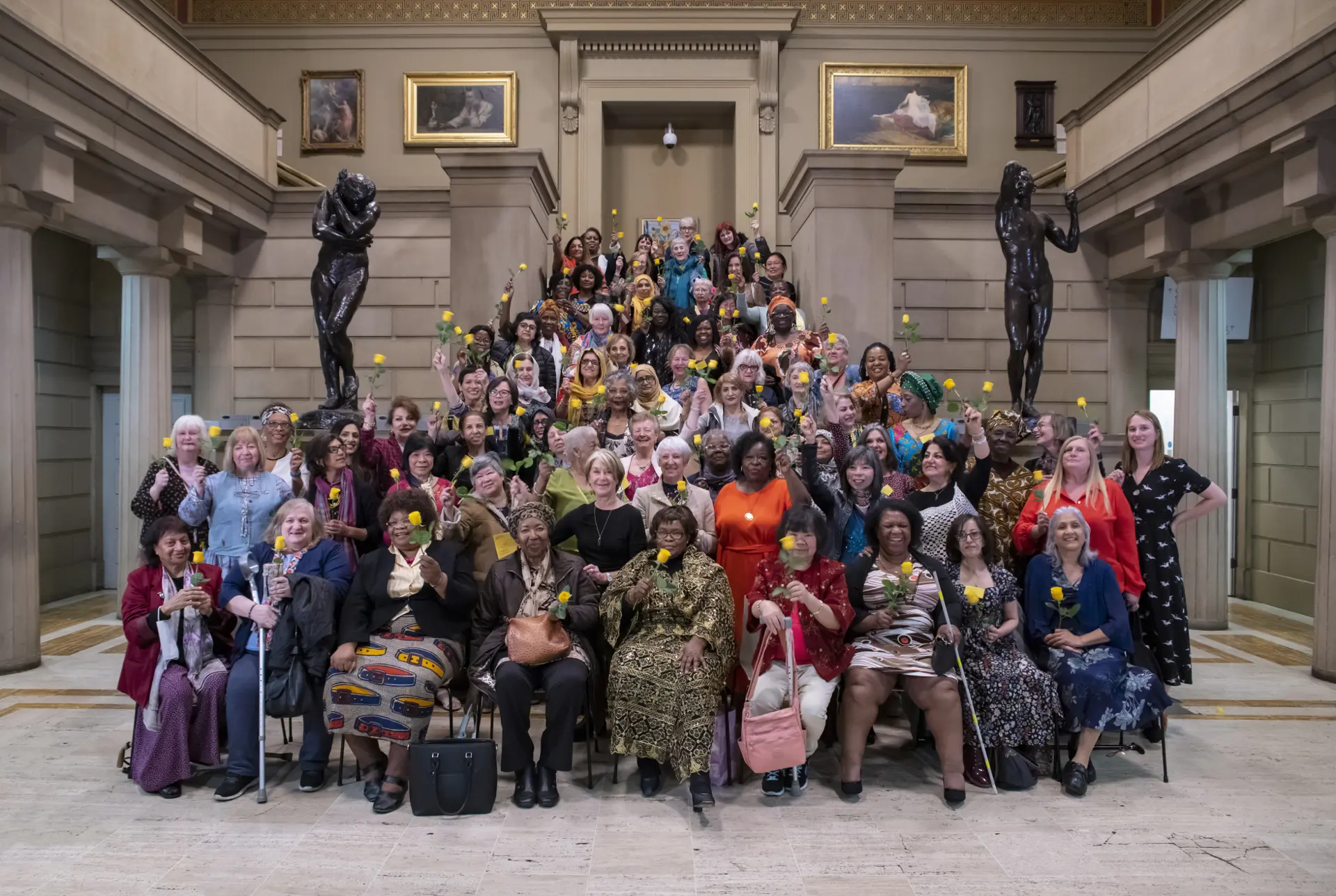 More than a hundred women holding yellow roses on the steps of a grand hall